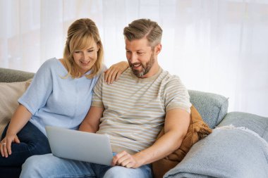 Orta yaşlı çift evdeki kanepede dizüstü bilgisayar kullanıyor. Sarı saçlı kadın ve gri saçlı adam internette geziniyor.. 