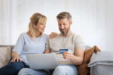 Orta yaşlı çift evdeki kanepede dizüstü bilgisayar kullanıyor. Orta yaşlı mutlu bir çiftin evde dizüstü bilgisayarla kredi kartı ödemesi çekimleri. 