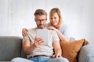 Orta yaşlı bir çift evdeki kanepede dokunmatik ped kullanıyor. Sarı saçlı kadın ve ağda gezinen gri saçlı adam.. 