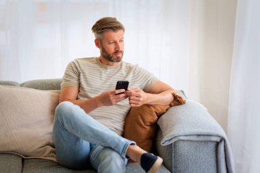 Orta yaşlı bir adam evdeki kanepede oturuyor ve akıllı telefonunu kullanıyor. Beyaz erkek mesajlaşmasıName. 