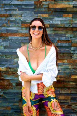 Duvarda dinlenirken güneş gözlüğü takan ve plaj kıyafeti giyen mutlu, uzun saçlı bir kadının portresi..