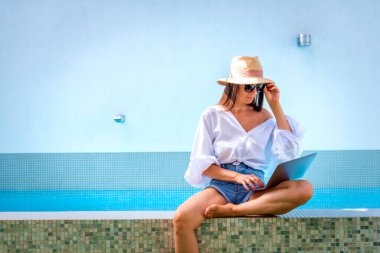 Havuzun kenarında oturan ve dizüstü bilgisayar kullanan bir kadın. Esmer kadın hasır şapka takıyor, günlük kıyafetler giyiyor ve internette kaşlarını çatıyor.. 