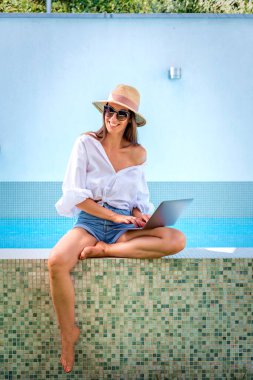 Havuzun kenarında oturan ve dizüstü bilgisayar kullanan bir kadın. Esmer kadın hasır şapka takıyor, günlük kıyafetler giyiyor ve internette kaşlarını çatıyor.. 