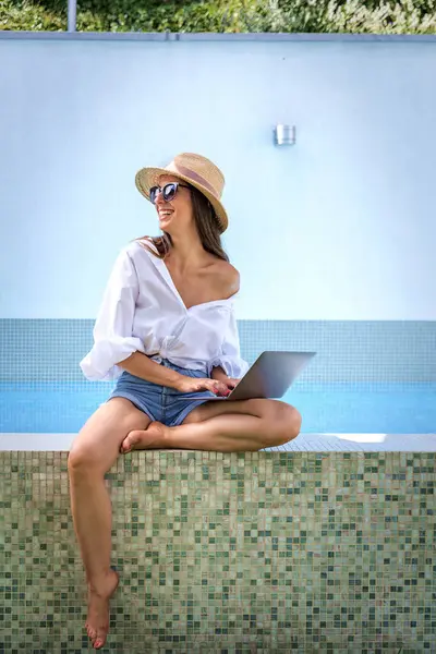 Havuzun kenarında oturan ve dizüstü bilgisayar kullanan bir kadın. Esmer kadın hasır şapka takıyor, günlük kıyafetler giyiyor ve internette kaşlarını çatıyor.. 