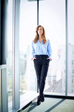 Modern bir ofiste yönetici bir iş kadınının portresi. Profesyonel kadın iş kıyafeti giyiyor ve düşünceli görünüyor. Tam boy çekim. 