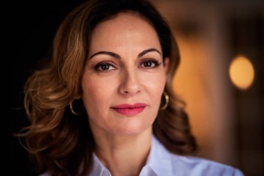 Kendinden emin bir kadın sıcak bir şekilde gülümsüyor, evin içinde ayakta duruyor. Sarı saçlı, beyaz bluz ve kırmızı ruj giyen bir kadın.. 