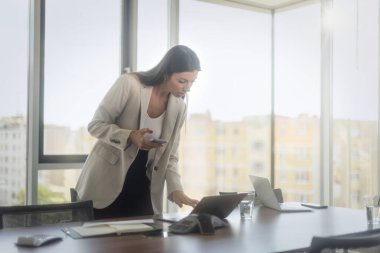 Ofisteki bilgisayarını ve akıllı telefonunu kullanarak gülümseyen bir iş kadını. Modern bir ofiste oturan çekici, profesyonel bir kadın..