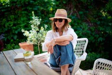 Sarı saçlı kadın arka bahçede oturuyor ve dinleniyor. Beyaz gömlek ve güneş şapkası giyen çekici olgun bir kadın.. 