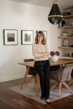 Modern evinde duran ve gülümseyen güzel bir kadının portresi. Orta yaşlı kadın gündelik giysiler giyiyor ve masa başında duruyor.. 