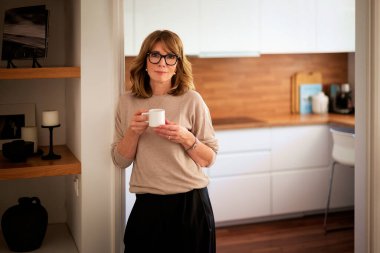 Gülümseyen orta yaşlı sarışın bir kadının portresi. Mutfakta tek başına dikilirken bir fincan kahve içiyor. Gündelik kıyafetler ve gözlük takan kendinden emin bir kadın.. 