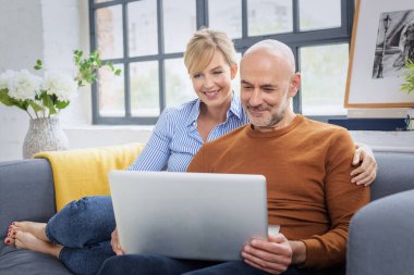 Evde dizüstü bilgisayar kullanan sevimli, olgun bir çiftin fotoğrafı. Sarı saçlı kadın ve sıradan kıyafetler giyen yakışıklı adam.. 