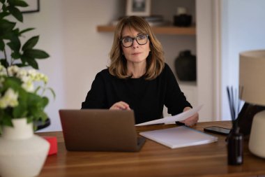 Evde dizüstü bilgisayarının önünde oturan sarı saçlı bir kadın. Evden çalışan orta yaşlı beyaz bir kadın. Merkez Ofis. 