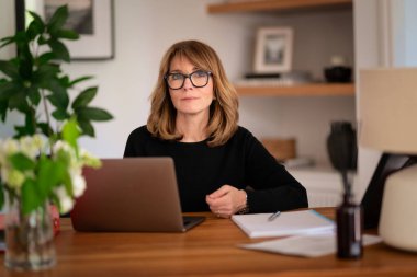 Evde dizüstü bilgisayarının önünde oturan sarı saçlı bir kadın. Evden çalışan orta yaşlı beyaz bir kadın. Merkez Ofis. 