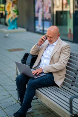 Şehirdeki bir bankta oturan yakışıklı iş adamı cep telefonu ve bilgisayar laptopu kullanıyor. Profesyonel erkek iş kıyafeti giyiyor ve çalışıyor. Tam menzil.. 