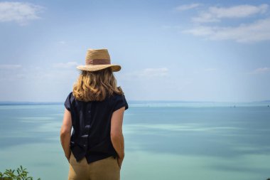 Sahilde durmuş hasır şapka takan ve deniz manzarasının tadını çıkaran bir kadın. Arka görünüm. 