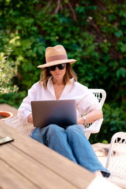 Arka bahçede masada otururken dizüstü bilgisayar kullanan sarı saçlı kadın. Evde çalışan çekici olgun bir kadın. Merkez Ofis. 