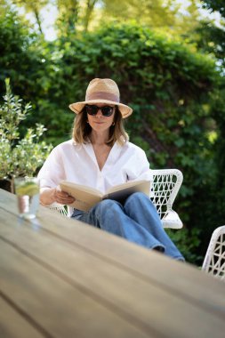 Sarışın kadın arka bahçede oturuyor ve kitap okuyor. Beyaz gömlek ve güneş şapkası giyen çekici olgun bir kadın. Evde dinlenen bir kadın. Tam boy çekim. 