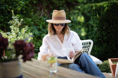 Sarışın kadın arka bahçede oturuyor ve kitap okuyor. Beyaz gömlek ve güneş şapkası giyen çekici olgun bir kadın. Evde dinlenen bir kadın. Tam boy çekim. 