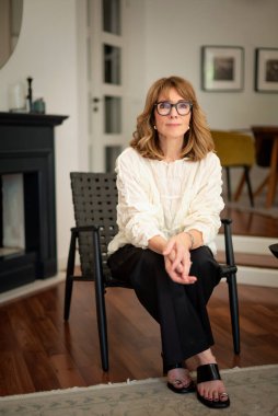 Güzel, orta yaşlı bir kadının rahat evinde koltukta oturduğu uzun bir çekim. Sarı saçlı kadın gömlek ve pantolon giyiyor.. 