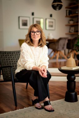 Güzel, orta yaşlı bir kadının rahat evinde koltukta oturduğu uzun bir çekim. Sarı saçlı kadın gömlek ve pantolon giyiyor.. 