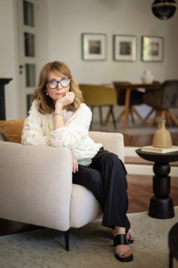 Güzel, orta yaşlı bir kadının rahat evinde koltukta oturduğu uzun bir çekim. Sarı saçlı kadın gömlek ve pantolon giyiyor.. 