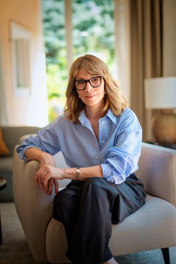 Modern evinde rahat bir koltukta oturan orta yaşlı güzel bir kadının fotoğrafı. Kendinden emin kadın gömleği ve pantolonu var ve kameraya gülümsüyor..  