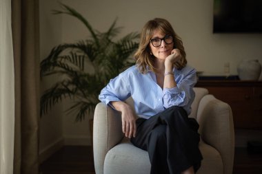 Modern evinde rahat bir koltukta oturan orta yaşlı güzel bir kadının fotoğrafı. Kendinden emin kadın gömleği ve pantolonu var ve kameraya gülümsüyor..  