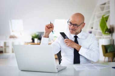 Ofis masasında dizüstü bilgisayarı olan beyaz bir adam oturuyor ve bir defter kullanıyor. Profesyonel iş adamı web konferansı yapıyor ve mutlu görünüyor.. 