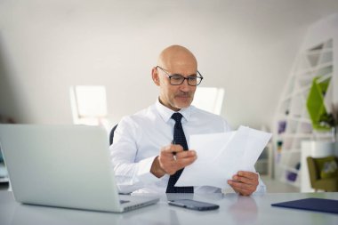 Ofis masasında dizüstü bilgisayarı olan beyaz bir adam oturuyor ve bir defter kullanıyor. Profesyonel işadamı dikkatli bakıyor ve elinde bir belge tutuyor..
