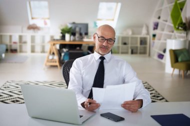 Ofis masasında dizüstü bilgisayarı olan beyaz bir adam oturuyor ve bir defter kullanıyor. Profesyonel işadamı dikkatli bakıyor ve elinde bir belge tutuyor..