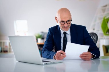 Ofis masasında dizüstü bilgisayarı olan beyaz bir adam oturuyor ve bir defter kullanıyor. Profesyonel işadamı dikkatli bakıyor ve elinde bir belge tutuyor..
