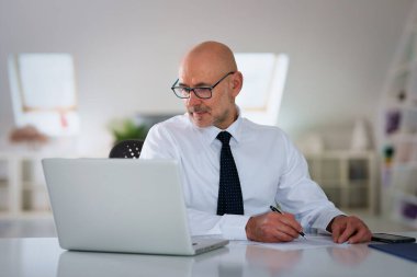 Ofis masasında dizüstü bilgisayarı olan beyaz bir adam oturuyor ve bir defter kullanıyor. Profesyonel işadamı düşünceli görünüyor ve web konferansı düzenliyor..