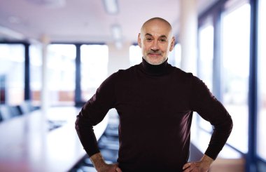 Toplantı odasında dikilip kameraya bakan kendinden emin orta yaşlı bir adam. Orta yaşlı erkek günlük kıyafetler giyiyor. Ellerini göğsüne koy.. 