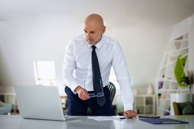 Ofis masasında dizüstü bilgisayarı olan beyaz bir adam duruyor ve bir defter kullanıyor. Profesyonel işadamı düşünceli görünüyor ve web konferansı düzenliyor..