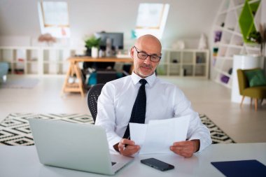 Ofis masasında dizüstü bilgisayarı olan beyaz bir adam oturuyor ve bir defter kullanıyor. Profesyonel işadamı dikkatli bakıyor ve elinde bir belge tutuyor..