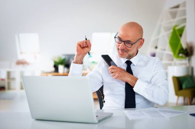 Ofis masasında dizüstü bilgisayarı olan beyaz bir adam oturuyor ve bir defter kullanıyor. Profesyonel iş adamı web konferansı yapıyor ve mutlu görünüyor.. 