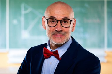 Orta yaşlı profesör matematik öğretiyor. Kendine güveni tam bir öğretmen. Tahtanın önünde durmuş kameraya bakıyor..