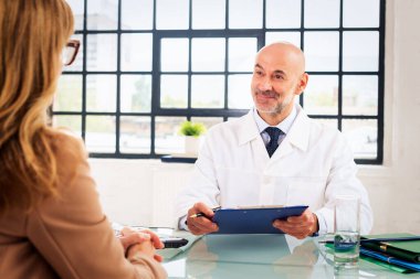 Masada oturan ve erkek doktoruyla kan testi sonuçları hakkında konuşan bir kadın. Erkek doktorun elinde bilgi panosu var ve bunu tartışıyorlar..