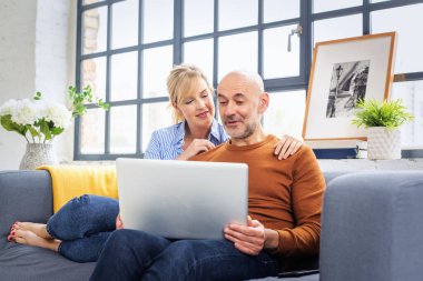 Evde dizüstü bilgisayar kullanan sevimli, olgun bir çiftin fotoğrafı. Sarı saçlı kadın ve sıradan kıyafetler giyen yakışıklı adam.. 