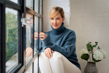 Çekici, sarı saçlı, pencerenin kenarında dinlenen bir kadının portresi. Siyah kazak ve beyaz pantolon giyen, derin düşüncelere dalmış kendinden emin bir kadın.. 