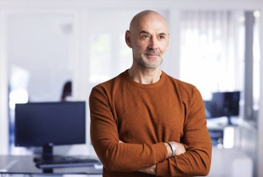 Ofiste duran orta yaşlı kel adam. Kendine güvenen iş adamı kazak giyiyor ve kameraya gülümsüyor..