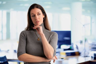 Çekici, orta yaşlı bir iş kadınının düşünceli bakışları. Esmer kadın balıkçı yaka kazak giyiyor ve ofiste dikiliyor. Boşluğu kopyala. 