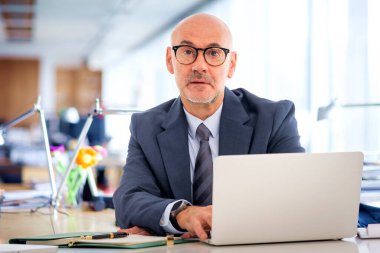 Takım elbiseli ve kravatlı orta yaşlı bir işadamı masada oturuyor ve kameraya bakıyor. Modern bir ofiste çalışan kendinden emin bir erkek.. 