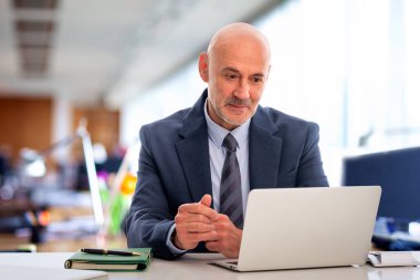 Orta yaşlı takım elbiseli ve kravatlı bir işadamı masada oturmuş bilgisayarının ekranına bakıyor. Modern ofis. Kendine güvenen profesyonel bir erkeğin video görüşmesi var.. 