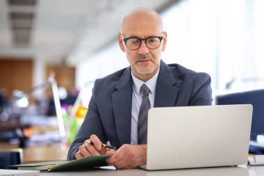Orta yaşlı takım elbiseli ve kravatlı bir işadamı masada oturmuş bilgisayarının ekranına bakıyor. Modern ofis. 