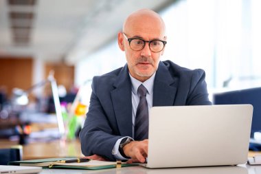Orta yaşlı takım elbiseli ve kravatlı bir işadamı masada oturmuş bilgisayarının ekranına bakıyor. Modern ofis. 