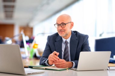 Orta yaşlı takım elbiseli ve kravatlı bir işadamı masada oturmuş bilgisayarının ekranına bakıyor. Modern ofis. 