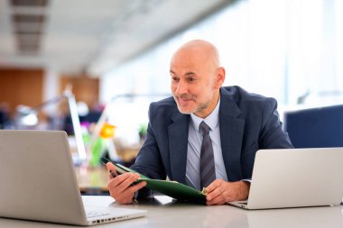 Orta yaşlı takım elbiseli ve kravatlı bir işadamı masada oturmuş bilgisayarının ekranına bakıyor. Modern ofis. 