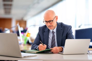 Takım elbiseli ve kravatlı orta yaşlı bir işadamı masada oturuyor ve not alıyor. Dizüstü bilgisayar kullanan profesyonel bir erkek. Ciddi yüz..