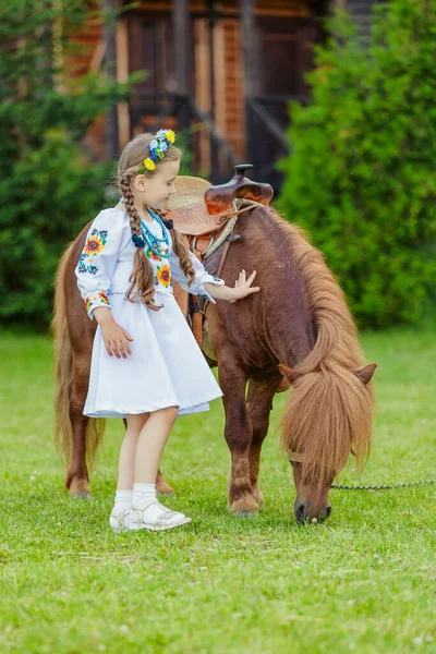 ウクライナの民族衣装の少女は芝生の上で草を食べるポニーをストロークします — ストック写真
